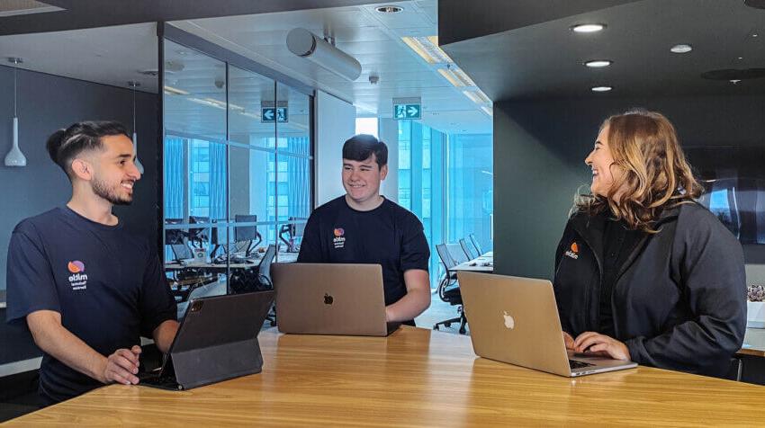 Three young people in Mitie apprentice-branded t-shirts laughing with each other. They stand around a wooden table in an office, using laptops.